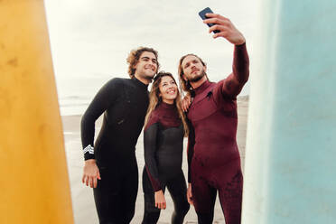 Gruppe glücklicher Surferfreunde in Neoprenanzügen, die in der Nähe von Surfbrettern stehen, während sie ein Selfie mit dem Smartphone am Strand während des Trainings machen - ADSF27723