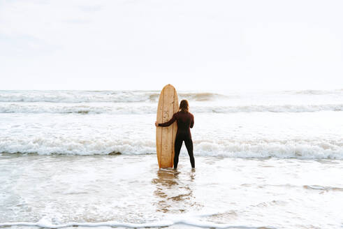 Rückenansicht eines nicht erkennbaren Surfers im Neoprenanzug, der mit seinem Surfbrett in Richtung Wasser schaut, um eine Welle am Strand bei Sonnenaufgang zu erwischen - ADSF27724