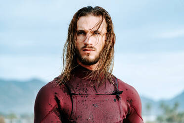 Junger gutaussehender Surfer mit langen Haaren im Neoprenanzug, der bei Sonnenaufgang am Strand steht und wegschaut - ADSF27725