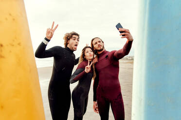 Gruppe glücklicher Surferfreunde in Neoprenanzügen, die in der Nähe von Surfbrettern stehen, während sie ein Selfie mit dem Smartphone am Strand während des Trainings machen - ADSF27726