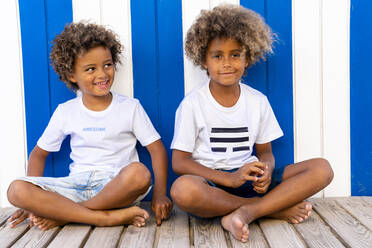 Smiling brothers sitting on wooden walkway against striped wall - DLTSF02043