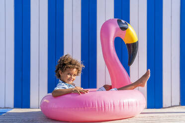 Niedlicher, lockig behaarter Junge sitzt in einem aufblasbaren Flamingo - DLTSF02053
