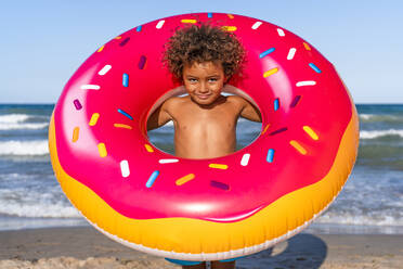 Junge ohne Hemd schaut durch aufblasbaren Donut am Strand - DLTSF02058