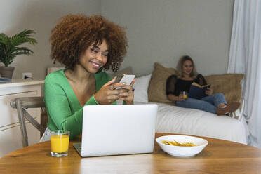 Smiling Afro businesswoman using smart phone at desk in home office - PNAF02060