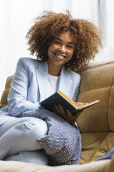 Lächelnde weibliche Fachkraft mit Buch auf dem Sofa zu Hause - PNAF02084