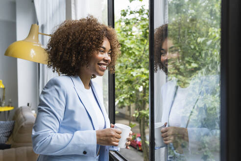 Glückliche junge Geschäftsfrau mit Kaffeetasse, die durch das Fenster ihres Hauses schaut - PNAF02102