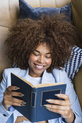 Lächelnde junge Geschäftsfrau im Blazer, die auf dem Sofa liegend ein Buch liest - PNAF02107