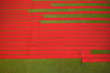 Aerial view of two people working in a field stretching red cotton fabric rolls in Narsingdi, Dhaka, Bangladesh. - AAEF10699