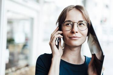 Female freelancer looking away while talking on smart phone and digital tablet - JOSEF05253