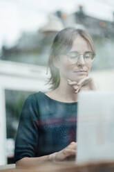 Female freelancer sitting with hand on chin using laptop in cafe seen through glass window - JOSEF05259