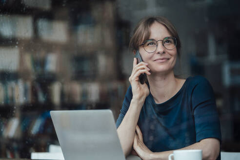 Lächelnde weibliche Fachkraft, die wegschaut und mit einem Smartphone in einem Café spricht, gesehen durch ein Glasfenster - JOSEF05266