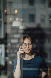 Young businesswoman talking on mobile phone seen through glass window - JOSEF05267