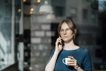 Junge Frau hält Kaffeetasse und spricht mit Smartphone - JOSEF05278