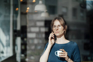 Young woman holding coffee cup talking on smart phone - JOSEF05278