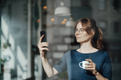 Junge Frau hält eine Kaffeetasse und schaut auf ihr Mobiltelefon - JOSEF05279
