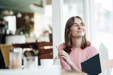 Lächelnde weibliche Fachkraft, die mit einem digitalen Tablet in einem Café sitzt und wegschaut - JOSEF05286
