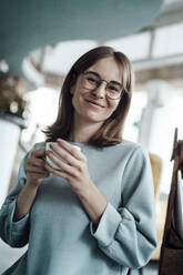 Lächelnde junge Frau mit braunen Haaren hält Kaffeetasse in Café - JOSEF05302