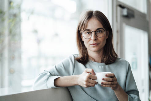 Junge Frau mit braunen Haaren hält eine Kaffeetasse, während sie in einem Café sitzt - JOSEF05306