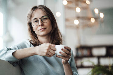 Young woman wearing eyeglasses holding coffee cup while sitting in cafe - JOSEF05307