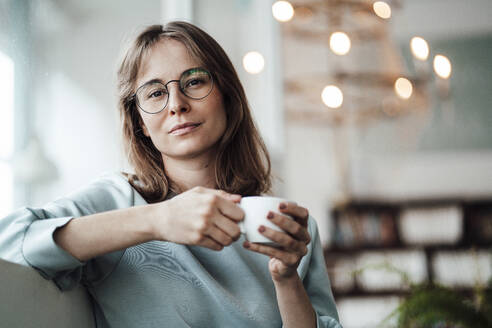 Junge Frau mit Brille hält eine Kaffeetasse, während sie in einem Café sitzt - JOSEF05307