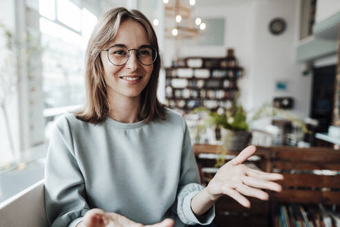 Lächelnde junge Frau mit Brille, die in einem Café sitzt und gestikuliert - JOSEF05310