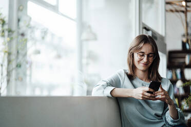 Smiling woman text messaging through smart phone sitting in cafe - JOSEF05315