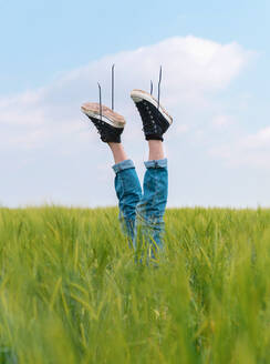 Umgekehrte gesichtslose Person in Jeans und schwarzen Turnschuhen, die ihre Beine aus dem grünen Gras eines Feldes im Sommer hebt - ADSF27742