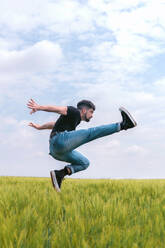 Side view man in denim jump with raised leg above tall grass in gloomy field - ADSF27743