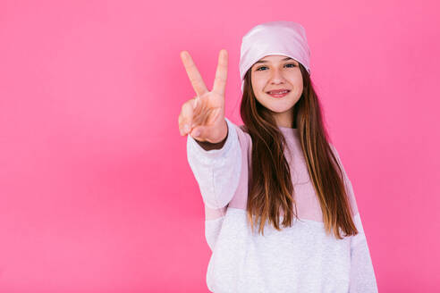 Young content female adolescent in headscarf for cancer awareness with outstretched arm demonstrating peace sign while looking at camera - ADSF27745