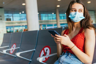 Weibliche Touristin mit Schutzmaske sitzt in der Abflughalle des Flughafens und wartet auf ihren Flug während der Coronavirus-Epidemie, während sie ihr Smartphone benutzt - ADSF27758
