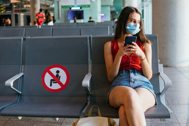 Female tourist wearing protective mask sitting in departure lounge of airport and waiting for flight during coronavirus epidemic while using smartphone - ADSF27759