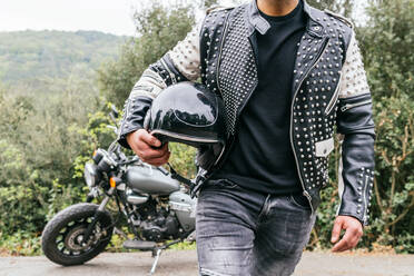 Anonymer männlicher Motorradfahrer in Jeans und Lederjacke, der einen Helm in der Hand hält, während er auf einer asphaltierten Straße neben einem geparkten modernen Motorrad steht - ADSF27763