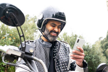 Bearded male biker in trendy leather jacket with rivets and protective helmet browsing phone while sitting on motorcycle parked near lush green forest - ADSF27765