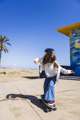 Aktive Frau in Freizeitkleidung fährt Skateboard auf der Straße entlang des Sandstrandes und der hohen Palmen während des Trainings und schaut weg - ADSF27769
