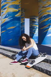 Full body side view of female in casual clothes sitting on stairs near skateboard browsing on smartphone on sunny street during training - ADSF27771