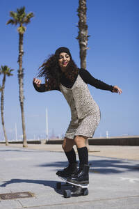 Ganzkörperansicht einer aktiven, fröhlichen Frau in einem Kleid, die auf einem Skateboard auf einer Straße entlang eines Sandstrandes mit hohen Palmen während des Trainings fährt - ADSF27773