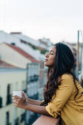 Side view of young Hispanic woman with closed eyes enjoying hot beverage while resting on balcony in evening - ADSF27778