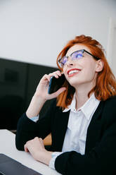 Junge Unternehmerin in formeller Kleidung, die mit einem Handy telefoniert, während sie zu Hause am Schreibtisch mit einem Laptop arbeitet - ADSF27810