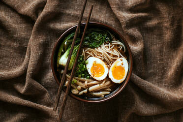 Top view of ceramic bowl with delicious ramen and chopsticks placed on table covered with brown tablecloth - ADSF27834