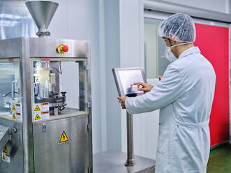 Seitenansicht eines männlichen Chemikers in Uniform bei der Bedienung einer Kapselfüllmaschine in einer pharmazeutischen Produktionsanlage - ADSF27845