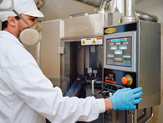 Seitenansicht eines männlichen Chemikers in Uniform bei der Bedienung einer Kapselfüllmaschine in einer pharmazeutischen Produktionsanlage - ADSF27852