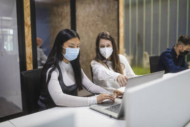 Multiracial female colleagues wearing masks sitting at table with laptop and discussing business project while working in coworking space - ADSF27854