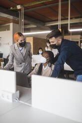 Group of diverse coworkers in protective masks gathering on laptop and discussing project while working together in contemporary workspace - ADSF27892