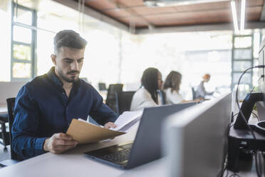 Concentrated male employee sitting at table with laptop and reading documents while working in spacious office with blurred colleagues - ADSF27896