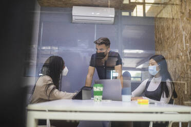 Through glass of company of multiracial coworkers in protective masks gathering at table and discussing project during business meeting - ADSF27898