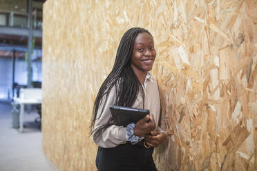 Lächelnde afroamerikanische Unternehmerin, die mit einem Tablet an der Wand eines Coworking Space steht und in die Kamera schaut - ADSF27907