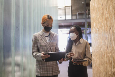 Multiethnic female colleagues with tablets and in protective masks standing in coworking space and discussing project - ADSF27909