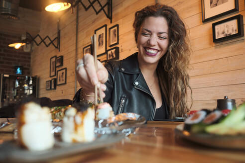 Smiling female eating tasty sushi in Japanese restaurant while sitting at wooden table - ADSF27925