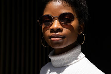 Junge afroamerikanische Frau in modischem Pullover und Sonnenbrille schaut in die Kamera, während sie in hellem Sonnenlicht vor schwarzem Hintergrund steht - ADSF27932
