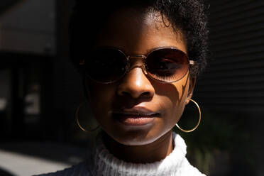 Junge afroamerikanische Frau in modischem Pullover und Sonnenbrille schaut in die Kamera, während sie in hellem Sonnenlicht vor schwarzem Hintergrund steht - ADSF27933
